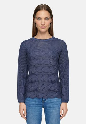 Femme aux cheveux bruns lisses portant un pull en maille torsadée bleu marine et un jean bleu, debout devant un fond clair uni.