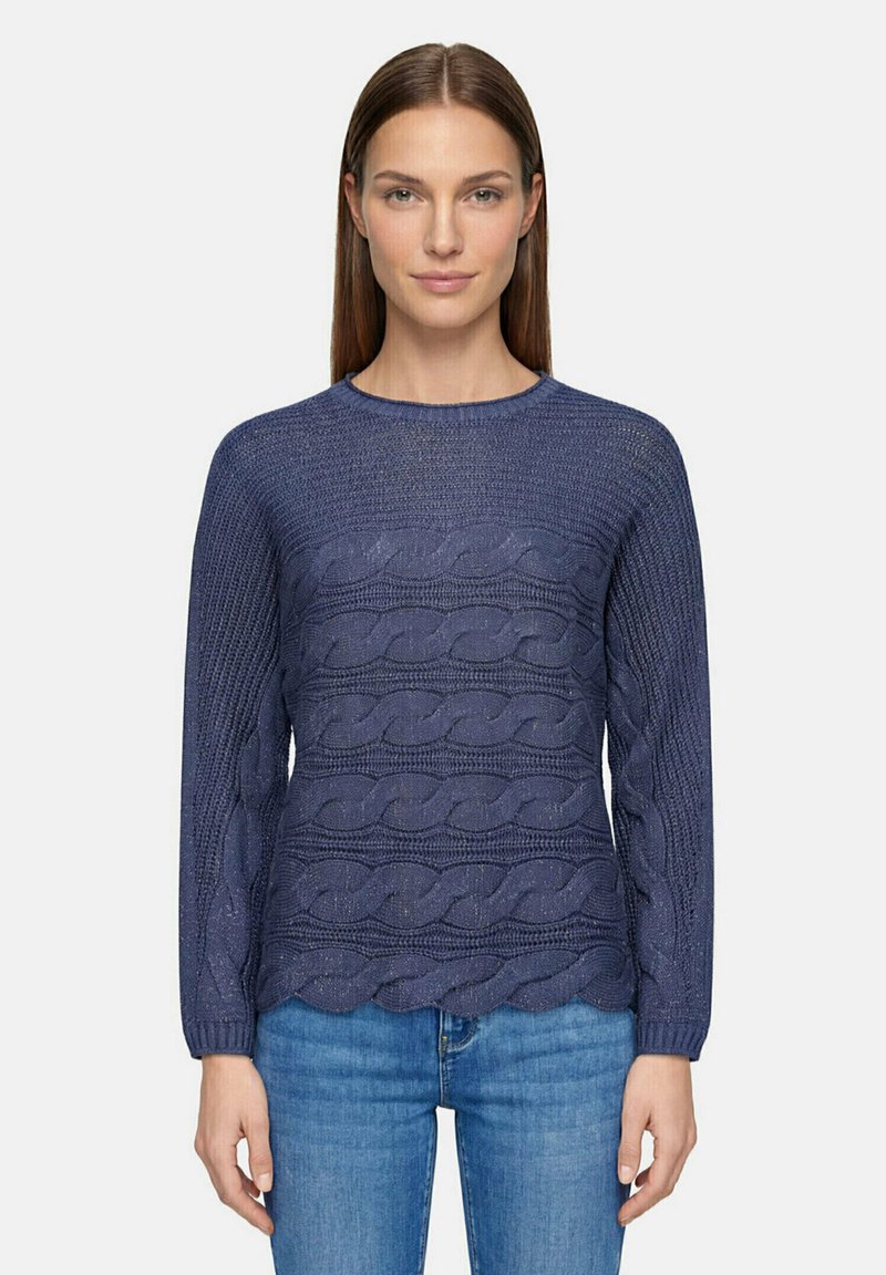 Femme aux cheveux bruns lisses portant un pull en maille torsadée bleu marine et un jean bleu, debout devant un fond clair uni.