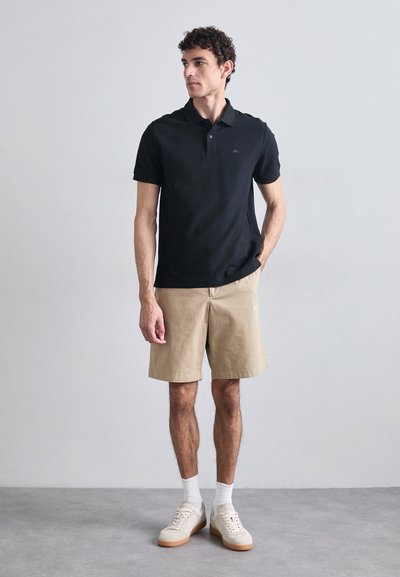 Camisa polo negra de manga corta con cuello, combinada con pantalones cortos beige. El modelo lleva zapatillas claras y calcetines blancos, de pie contra un fondo liso.