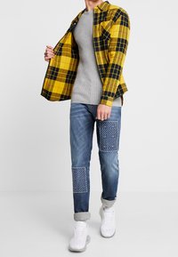 Żółto-czarna koszula w kratę nałożona na szary, prążkowany sweter; niebieskie dżinsy z niebieskimi łatami w wzory; białe trampki.