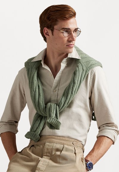 Light green cable-knit sweater draped over a cream collared shirt, paired with light beige trousers, showcasing round glasses and a blue watch.