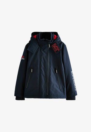 Marineblauwe geïsoleerde jas met rode accenten, uitgerust met ritszakken, gebreide manchetten en merknaam op de mouw en kraag.