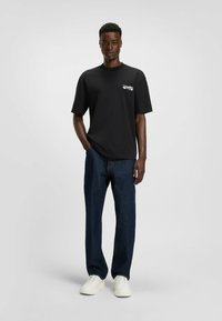 Sort bomulds T-shirt med korte ærmer og et hvidt logo på brystet, parret med mørkeblå denimjeans og hvide sneakers.