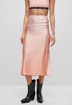 Femme portant une jupe midi en satin rose avec un détail à lacets sur le côté, un top court et des bottes de cow-boy noires mi-mollet, debout devant un fond uni.