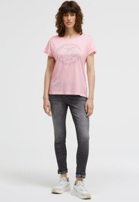 Light pink short-sleeve t-shirt featuring a circular graphic text design. Paired with dark gray fitted jeans and white sneakers.