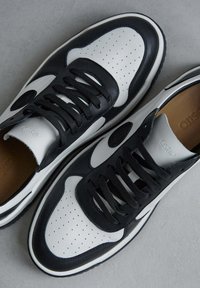 Černobílé tenisky s perforovaným koženým svrškem, černými šňůrkami, polstrovaným límcem a kontrastní podrážkou. Logo značky na jazyku.