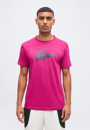 Junger Mann mit kurzen Haaren, der ein knallpinkes T-Shirt mit einer großen dunklen Krokodil-Grafik und weiße Shorts mit schwarzen Details trägt.