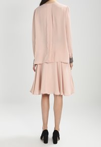 Abito rosa pallido con gonna fluida e maniche lunghe. I polsini dettagliati presentano inserti di pizzo scuro. Indossato con scarpe con tacco alto nere.