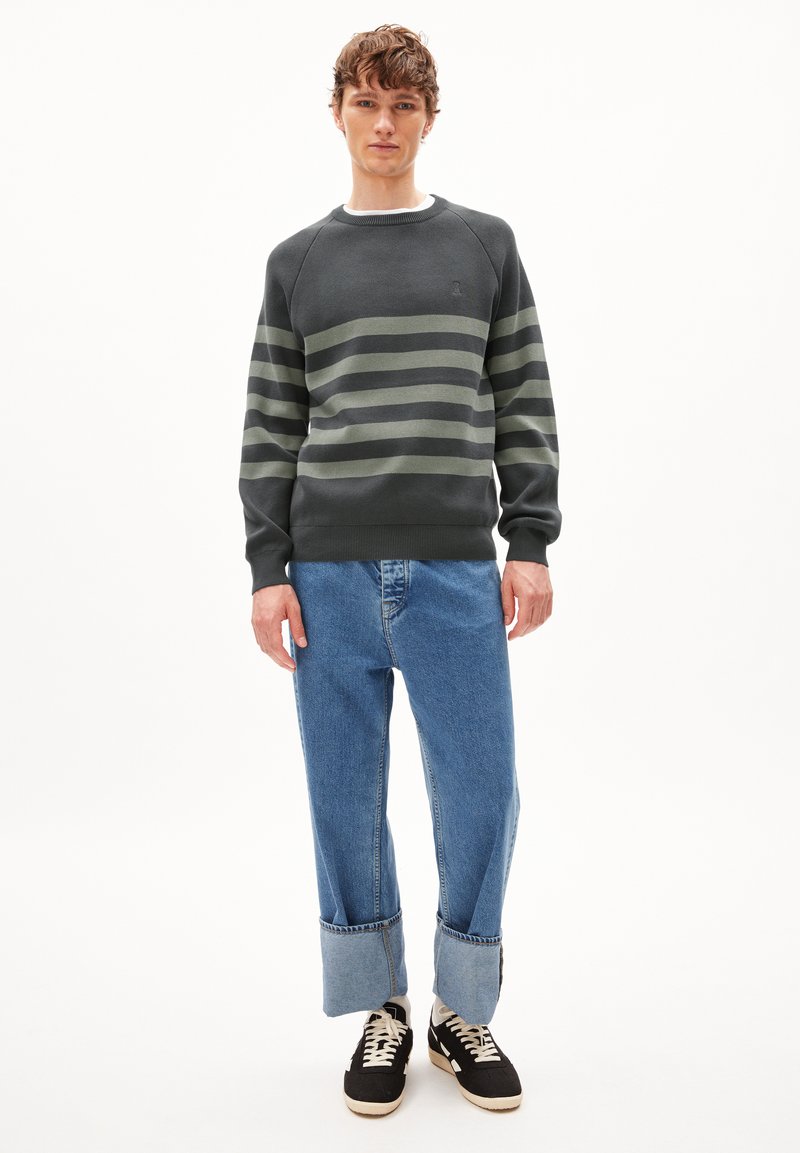 Dunkelgrauer Pullover mit grünen horizontalen Streifen, rundem Halsausschnitt und gerippten Bündchen, kombiniert mit hellblauen weit geschnittenen Jeans und schwarzen Sneakern.