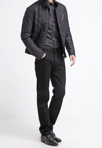 Svart vadderad jacka med dragkedja och bröstficka, lagd över en mörk knapp-up skjorta och svarta byxor, tillsammans med svarta skor.