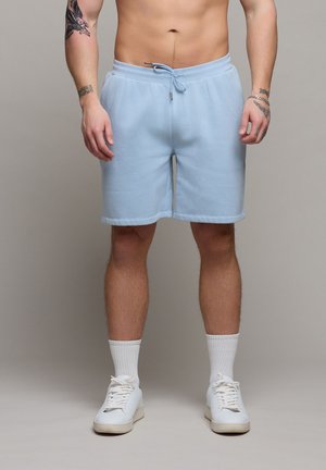 Männlich trägt hellblaue Shorts mit Kordelzug, weiße Kniestrümpfe und weiße Turnschuhe, sichtbar tätowierte Arme und oberkörperfrei.