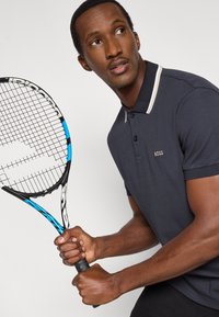 Μαύρο πόλο μπλουζάκι με αντίθετο γιακά, με λογότυπο "BOSS"· κρατώντας μια μαύρη και μπλε ρακέτα τένις με υφή λαβής.