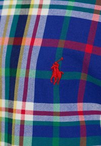 Rutig skjorta med blå bakgrund och röda, gröna och vita linjer. Den har en broderad röd polo-spelare logotyp på bröstet.