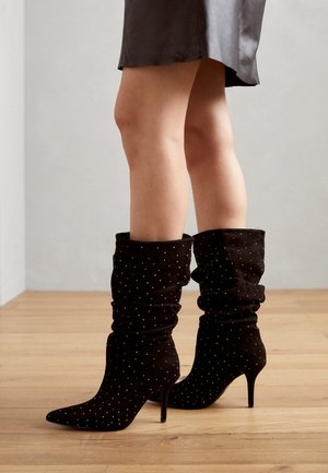 Gambe con stivali scamosciati neri a metà polpaccio con tacchi a punta e piccole borchie d'argento, poggiati su pavimento di legno.