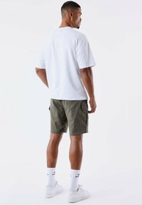 Camiseta blanca de manga corta, pantalones cortos tipo cargo en verde oliva y zapatillas blancas, con calcetines blancos que tienen un logo negro. Diseño simple y casual.