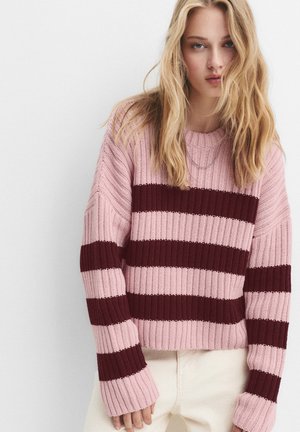 Jonge vrouw met lang blond haar draagt een gebreide trui met roze en donkerrode strepen en lichte broek tegen een effen witte achtergrond.