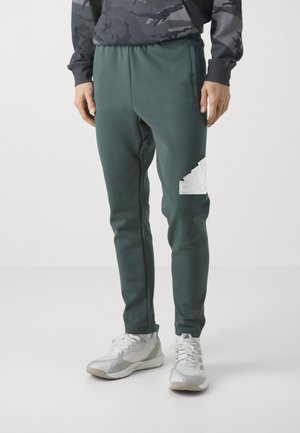 Pantalones de chándal verdes hechos de tejido suave, con una cinturilla elástica y un logo blanco de Adidas en el muslo izquierdo. Combínalos con zapatillas ligeras.