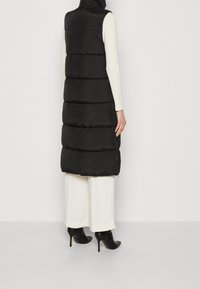 Svart pufferväst med hög krage, quiltad design och sidodragkedja. Visas med en vit långärmad tröja och vida vita byxor.