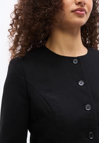 Γυναίκα με κατσαρά μαλλιά που φοράει μαύρο σακάκι με κουμπιά και στρογγυλό λαιμόκοψη, μπροστά σε απλό φόντο.