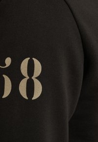 Sudadera negra con textura acanalada; presenta un gran número "58" en color blanco roto impreso en la manga izquierda. Diseño simple y moderno.