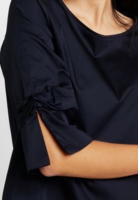 Marinblus i mjukt tyg med rund halsringning och korta ärmar prydda med knytningar vid ärmsluten.