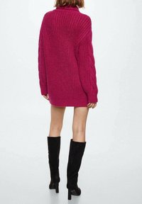 Femme portant un long pull texturé magenta et des bottes à talons hautes noires jusqu'aux genoux, debout de dos devant un fond uni.