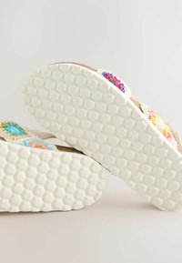 Platta sandaler med en strukturerad vit gummisula. Remmarna har färgglada, blommiga virkade mönster på en bakgrund av naturlig fiber.