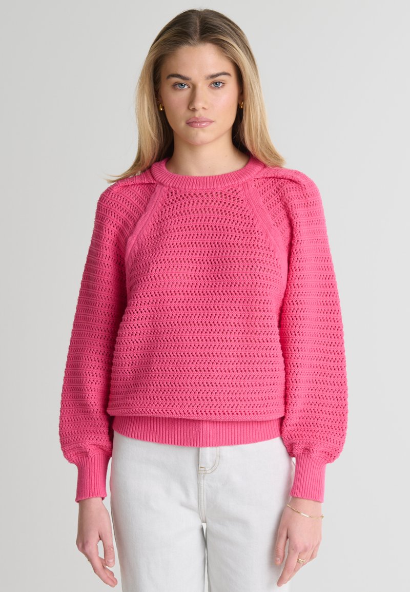 Różowy sweter z dzianiny o fakturowanym wzorze, z ściągaczami przy mankietach i u dołu, w luźnym fasonie. Posiada rękawy raglanowe i okrągły dekolt.