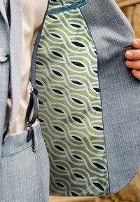 Mann im grauen Anzug zeigt das Innenfutter seiner Jacke mit grünem und schwarzem geometrischem Kettenmuster.