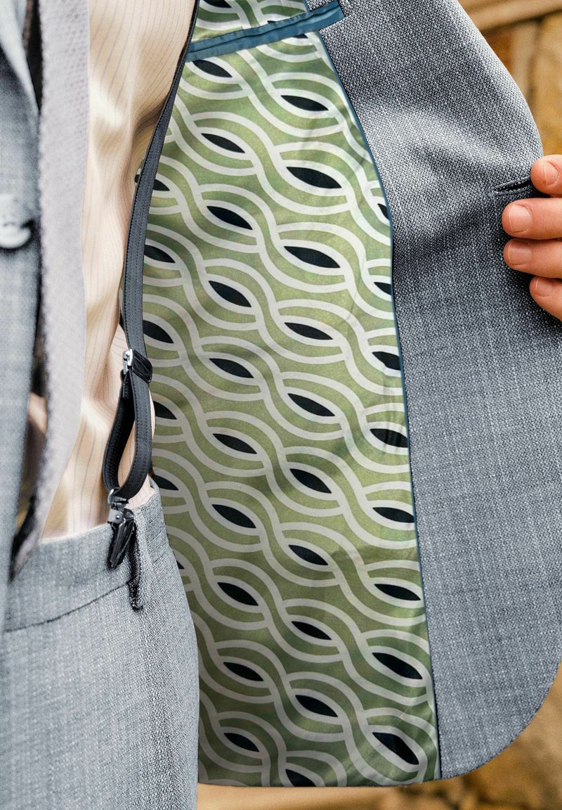 Mann im grauen Anzug zeigt das Innenfutter seiner Jacke mit grünem und schwarzem geometrischem Kettenmuster.