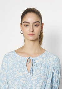 Blusa floral em azul claro com decote em gota, detalhes de ombro franzido e fecho com laço, feita de um tecido macio e texturizado.