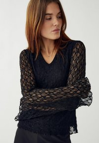 Čierna čipkovaná blúzka s dlhými priehľadnými rukávmi, výstrihom do V a textúrovanými kvetinovými vzormi. Mäkká tkanina s ľahkým, vrstveným vzhľadom.