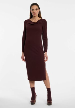 Woman standing wearing a fitted long-sleeve dark burgundy dress with a draped neckline and matching heeled sandals with socks.