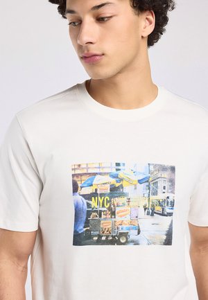 Jonge man draagt een wit T-shirt met een print van een hotdogkarretje in de straten van NYC met gele parasols en straatachtergrond.