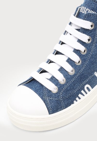 Gros plan d'une basket montante en denim bleu avec des lacets blancs, un embout en caoutchouc blanc et un texte blanc partiel sur le côté.