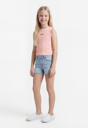 Κορίτσι που χαμογελά με μακριά ξανθά μαλλιά, φορώντας ροζ αμάνικο τοπ της Nike, ανοιχτό μπλε τζιν σορτς και λευκά slip-on αθλητικά παπούτσια.