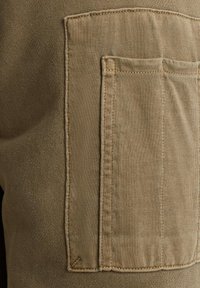 Khaki stof met geribbelde textuur heeft een grote zak op de zijkant, omlijnd met contrasterende stiksels.