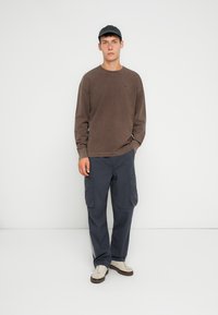 Hnedá košeľa s dlhým rukávom, námornícke cargo nohavice s vreckami, svetlé topánky a tmavá čiapka. Jednoduchý dizajn, uvoľnený strih a textúrovaná tkanina.