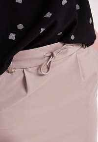 Pantalon beige avec taille à cordon, présentant des plis subtils et une texture lisse. Haut noir avec un motif géométrique en gris clair.