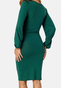 Robe verte à manches longues en tissu extensible. Silhouette ajustée avec des manches froncées et un léger ruissellement à la taille. Modèle mi-long.