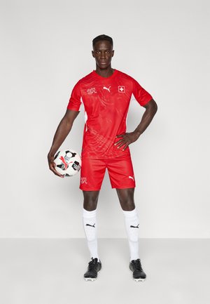 Piros focimez és rövidnadrág fehér díszítésekkel, logóval és struktúrált mintával. Fekete stoplis cipők és fehér zoknik logókkal. Egy focilabdát tart.