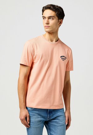 Lyserød kortærmet T-shirt lavet af bomuld, med rund hals og et lille marineblåt logo-design på venstre brystområde.