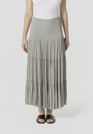 Žena oblečená v bielej tielkovej blúzke, svetlosivej viacvrstvovej maxi sukni so záhybmi v páse a čiernych sandáloch s otvorenou špičkou na jednoduchom pozadí.