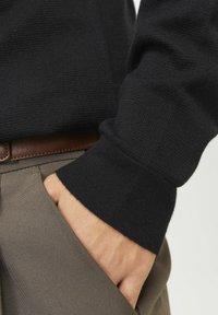 Melns adīts džemperis ar teksturētu apdari, ar pieguļošu aproci. Apvienots ar gaiši brūnām biksēm, roka ielikta kabatā.