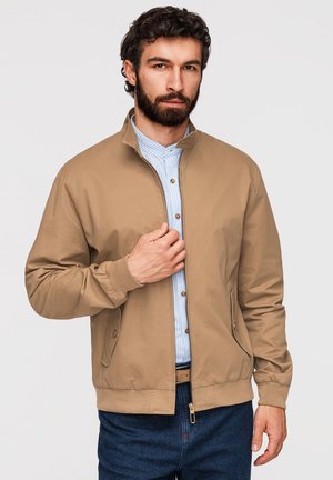 Homme aux cheveux bouclés foncés et à la barbe, portant une veste zippée beige par-dessus une chemise bleue à rayures boutonnée et un jean foncé.