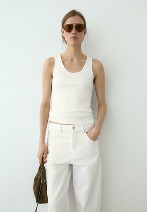 Femme portant un débardeur blanc et un pantalon blanc, lunettes de soleil marron, tenant un sac à main marron avec une main dans la poche sur fond blanc.