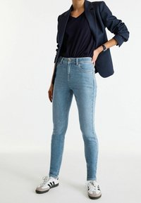 Vaqueros de mezclilla azul claro y ajustados con una cintura alta, complementados con un blazer negro y una blusa de pico en azul marino. Combinados con zapatillas negras y blancas.