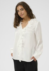 Biała bluzka z dekoltem w kształcie litery V, ozdobiona warstwowymi, płatkowymi detalami na przodzie oraz długimi rękawami. Gładka, lekka tkanina.