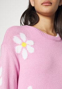 Rosa stickad tröja med stora vita blommor och gula mittdelar. Designen har en rund halsringning och ett texturerat stickmönster.