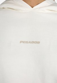 Biele mikina vyrobená z mäkkého materiálu, ktorá má predné klokanie vrecko a slovo "PEGADOR" v jemnom béžovom písmene nad vreckom.